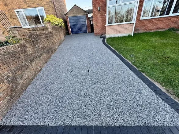 A grey resin bound driveway with a charcoal block paving border, running between a brick wall and a lawn up to a blue garage door.